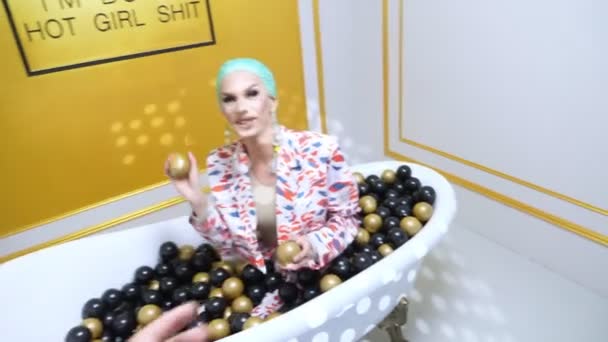 A handsome transgender guy in a blue wig and make-up sits in a bathroom with plastic balls and throws balls at a guy standing next to him. first-person view – Stock-video
