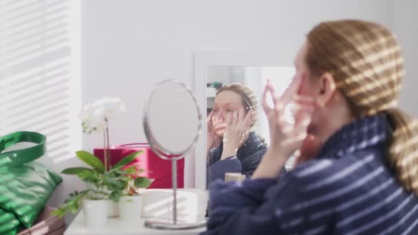 Жінка мазає обличчя кремом вдома, сидячи перед дзеркалом. тінь падає на неї з жалюзі — стокове відео