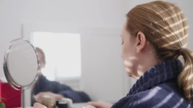 a woman smears her face with cream at home sitting in front of a mirror. a shadow falls on her from the blinds