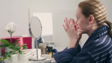 a woman smears her face with cream at home sitting in front of a mirror. a shadow falls on her from the blinds