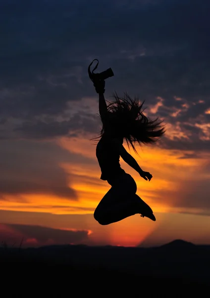 Girl silhouette jumping with photo camera in hand in sunset. Low key shot. — Stock Photo, Image
