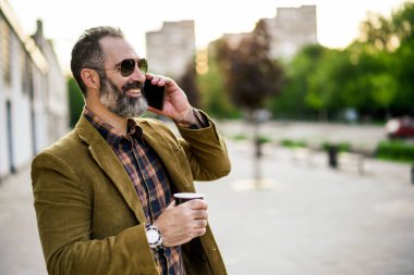 Sakallı modern iş adamının portresi. Şehir caddesinde dururken cep telefonu kullanıyor ve kahve içiyor..