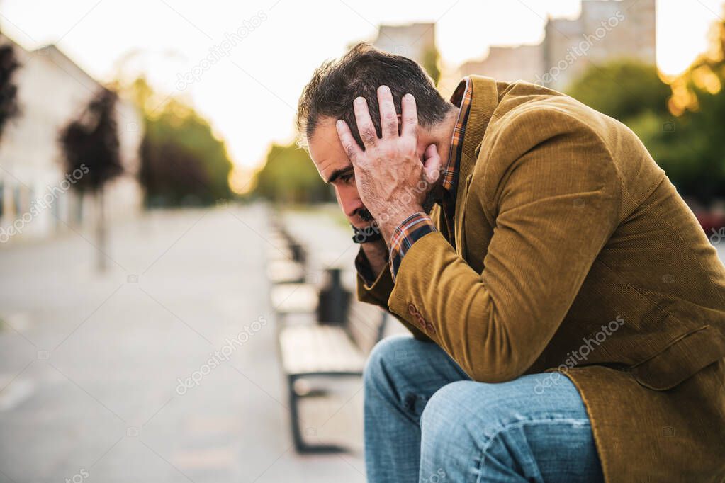 Depressed businessman having headache and sitting on a bench in the city street.