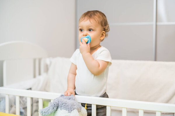 Image of cute baby boy with pacifier  spending time in crib.