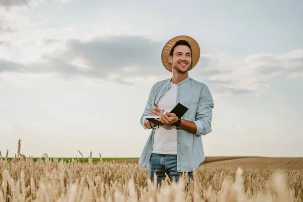 彼の成長する小麦畑に立っている間 幸せな農家はノートを書く — ストック写真