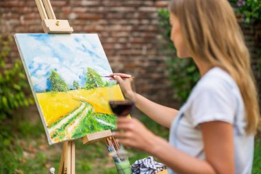 Woman enjoys  painting on canvas and drinking wine outdoor.
