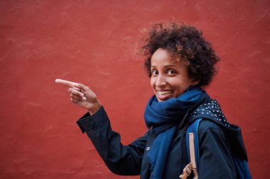 Gülümseyen siyahi bir kadının portresi kollarını gündelik kıyafetlerle gösteren ve kırmızı duvarda kısa kıvırcık saçlı bir kadın.