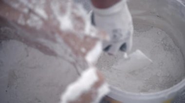 Koruyucu eldivenli işçiler kovaya beyaz alçı tozu dökerek solüsyonu inşaat alanındaki plastik kovada suyla karıştırıyorlar. Yeni macun duvarları için havan topu hazırlayan inşaatçı