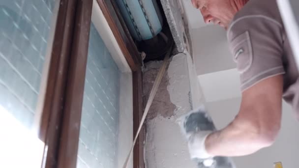 Low angle view of professional construction worker aligning and smoothing plastered wall with white finishing paste solution near the window with old external roller shutter blinds. Builder in — Stock Video