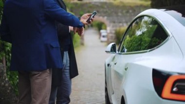 ROME, ITALY - 28 Nisan 2021: Roma 'da en kaliteli kiralık elektrikli araba, yüksek nitelikli işletme yöneticisi beyaz Tesla arabasının uzaktan kumandalı işlevlerini müşteriye gösteriyor, akıllı telefon uygulamaları kullanan adam