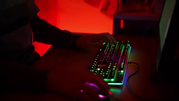 Jugador profesional sentado en la mesa en la habitación iluminada de color rojo oscuro y jugando en línea con el teclado y el ratón retroiluminados espectro, pirata informático que participa en torneos de vídeo ciberespacio con otros virtuales — Vídeo de stock