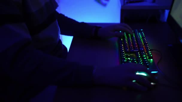 Темная комната освещена синими неоновыми огнями и ленивым человеком, играющим в онлайн видео игры день и ночь. Крупный план лиц, щелкающих пальцами на клавиатуре компьютера с подсветкой спектра. Видео — стоковое видео