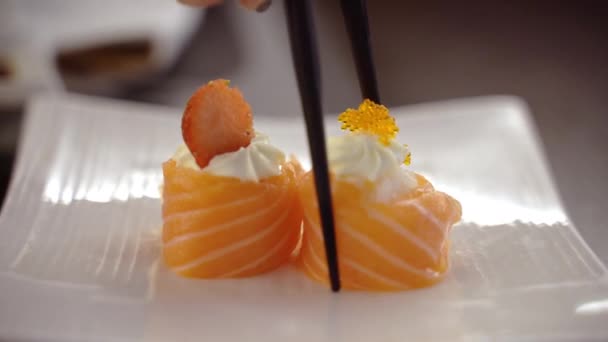 Mulher mão segurando pauzinhos e comer tradicional japonês sushi rolos no restaurante, dois japoneses sushi rolos com queijo Filadélfia, vermelho cru salmão fatias, fatia de morango e caviar amarelo como — Vídeo de Stock