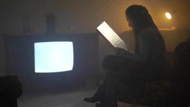 Сконцентрированная журналистка, читающая газету, сидящая на кресле в туманной комнате с темно-желтым освещением, женщина, сидящая перед большим винтажным телевизором с полосатым статическим шумом. Старое ретро — стоковое видео