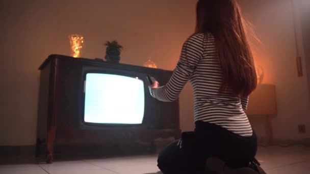 Filmový záběr tmavě oranžové osvětlené místnosti s retro nábytkem a blikající retro televizí, pohled na brunetku sedící na podlaze a měnící se kanály s dálkovým ovládáním — Stock video