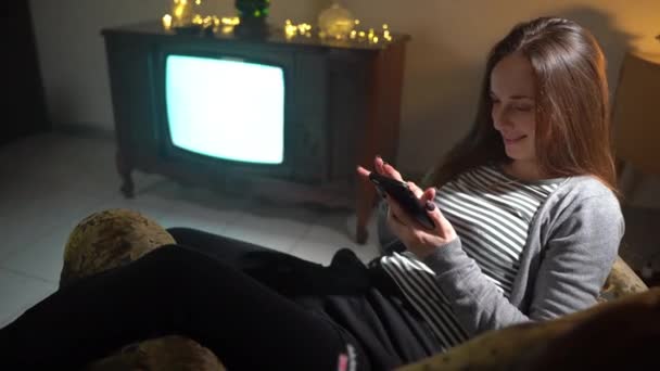 Mujer sonriente relajándose en casa en la habitación oscura con muebles vintage, mujer usando teléfono inteligente, charlando en línea o haciendo compras y sonriendo. Mujer sobre fondo de retro TV con pantalla parpadeante — Vídeos de Stock