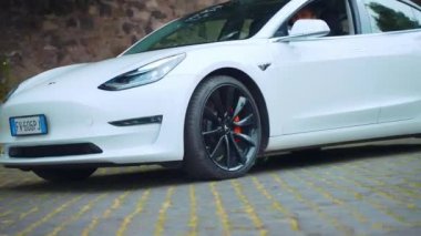 ROME, ITALY - 28 Nisan 2021: Roma 'nın kaldırım taşı caddelerinde beyaz yenilikçi Tesla araba test sürüşü direksiyonun başında yavaşça duran Tesla arabası, siyah tasarlanmış tekerleğin yakın görüntüsü