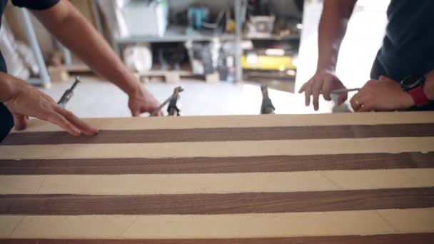 Kézművesek a műhelyben dolgoznak faanyagokkal, ácsok acélbilincseket használnak, hogy meghúzzák a ragasztott fa rudakat a munkapadon. Asztalos asztalosműhelyek fából készült remekművek készítéséhez — Stock videók