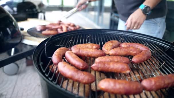 Juicy bratwurst sausages grilling on bbq grid for Oktoberfest party celebration in Germany. Barbecue lover using kitchen tongs for turning around german sausages roasting on charcoal and taking away — Stock Video