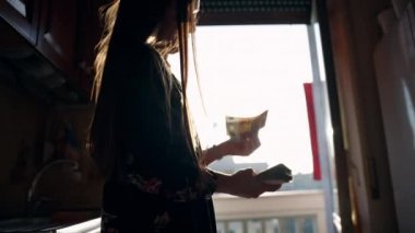 Evde para sayma süreci, kadın gelir ve gelir hesaplaması. Konsantre kadın, kontrastlı güneş ışığında kağıt para yığınından banknotlar sayıyor, para biriktirme kavramı
