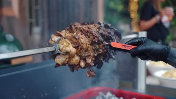 Person in black gloves shoving cooking thermometer inside the roasting pork meat for checking readiness, meat pieces rotating on barbecue skewer and roasting on burning fire and rising smoke. Barbecue — Stock Video