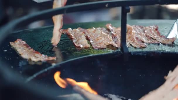 Orange and yellow fire flames on foreground, thin crispy bacon slices roasting on bbq grid for picnic party. Process of grilling juicy bacon slices, person using cooking tongs for turning around bacon — Stock Video