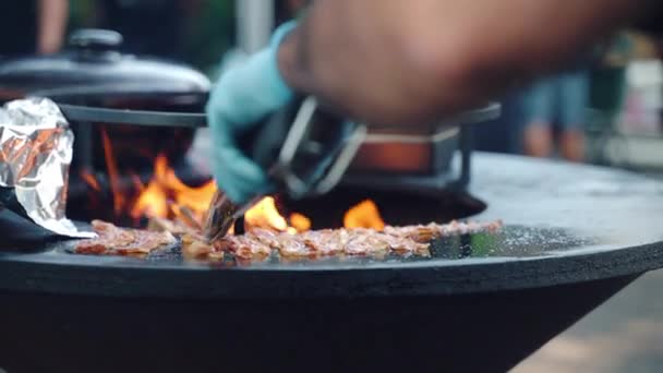 Festival de churrasco tradicional no fim de semana, chef qualificado em luvas usando pinças de cozinha para virar fatias de bacon suculentas grelhando em fumantes grelha de churrasco com fogo de laranja ardente — Vídeo de Stock