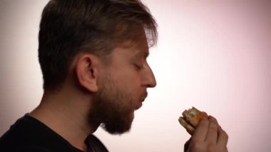 Zevkli hamburgerle yemek yiyen genç bir adamın profili, fast food restoranından alınmış et burgerli aç bir erkeğin lezzetli ekmeği. Sağlıklı genç adam evde sağlıksız fast-food yiyor, sorun bu.