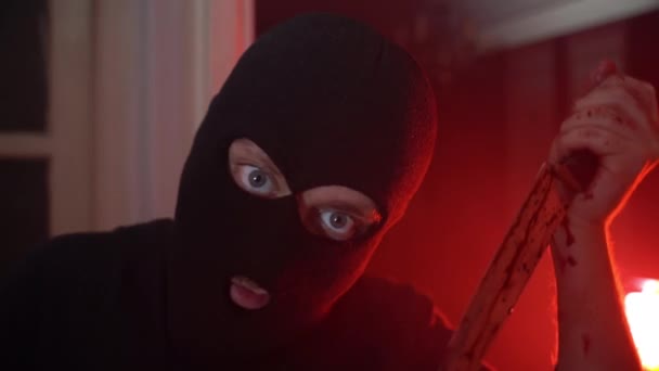 Portrait of spooky serial killer in black balaclava with demon eyes looking on camera and holding bloody knife in hands, psycho maniac ready for attacking victim with aggression and cold weapon. Cruel — Stock Video