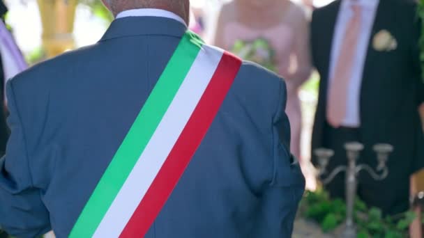 Zpět pohled na tradiční italský svatební úředník se třemi barvami vlajkovou stuhou přes rameno držící svatební řeč před novomanželským párem, krásný svatební obřad venkovní pod — Stock video