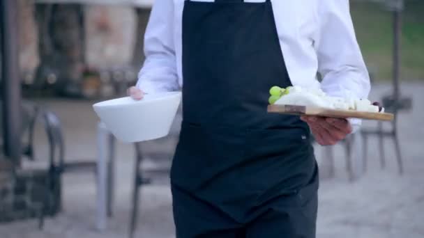 Официант ресторана в униформе, обслуживающий банкетный стол, украшенный наружным оформлением стола, слуга, держащий в руках белую миску с моцареллой и деревянную доску с ломтиками традиционного сыра. Кейтеринг — стоковое видео