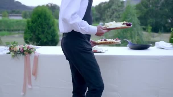 Kelner w mundurze restauracji serwujący luksusowe bankietowe party nakrywające do stołu na świeżym powietrzu, służący trzymający drewniane deski z plastrami różnych tradycyjnych rodzajów sera. Usługi cateringowe dla — Wideo stockowe
