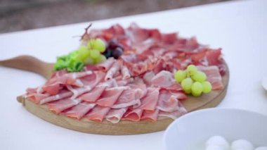 Yeşil ve siyah üzümlerle süslenmiş leziz mevsimsel prosciutto dilimleriyle özel İtalyan antipasto, ziyafet masasında taze mozzarella topları bulunan beyaz seramik kase. Yemek servisi.