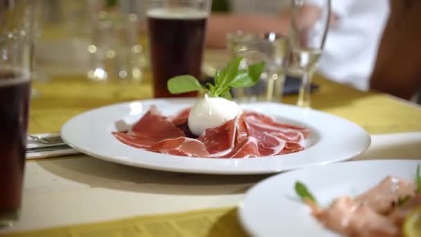 전통적 인 이탈리아 전통적 인 저녁 식사에서 점심이나 식사를 하고 있는 사람들이 있는 위에 신선 한 모차렐라와 바질 잎을 곁들인 흰 접시 사진을 찍는 여성 — 비디오