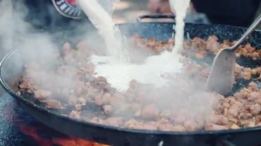 Yanan ateşte büyük metal bir kazan pirinç, kızarmış sosis ve tavuk parçaları ile sıcak bir kazan dökülüyor. Sokak yemekleri, kazanda yemek pişirme.