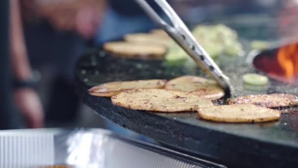 Chef utilisant des pinces de cuisson pour mettre des tranches de pommes de terre grillées et d'oignon prêtes dans un récipient en papier d'aluminium pour servir la table de pique-nique, tranches de légumes juteuses rôties sur barbecue grill de fumeur professionnel — Video