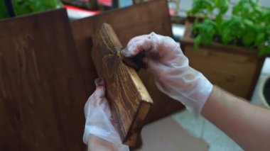 Professional carpenter working with wood in workshop outdoors, man in gloves coating wood board with brown colour protection using sponge. Joiner processing wood and staining surface to protect from
