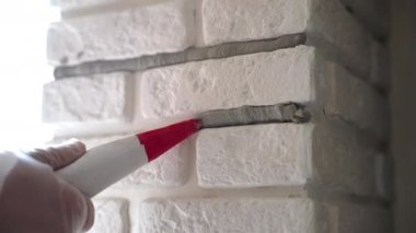 Workman in protective gloves filling joints with grout using construction syringe, builder finishing white decorative stones on interior wall. Applying mortar between stone plates, wall restoration