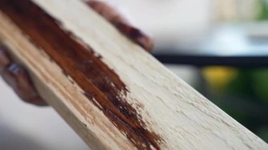 Woodman in transparent gloves moving along the wood plank surface with brown coloured sponge, carpenter covering wood surface with special colour protection, joiner varnishing hardwood in workshop