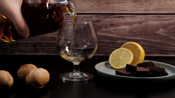 Handen häller konjak från flaskan i vinglaset. Bredvid den på en tallrik står en citron och chokladbitar — Stockvideo