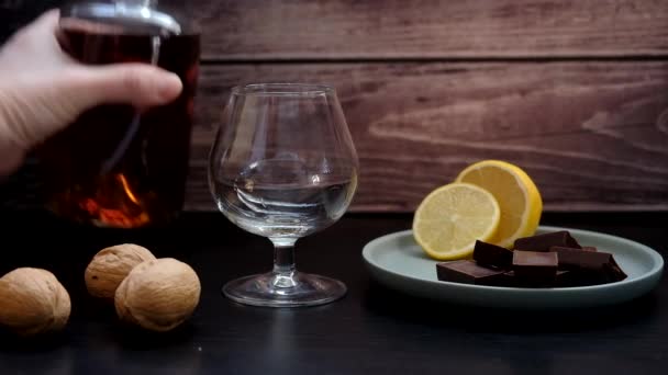 A kéz konyakot önt az üvegből a borospohárba. Mellette egy tányéron egy citrom és egy darab csokoládé. — Stock videók