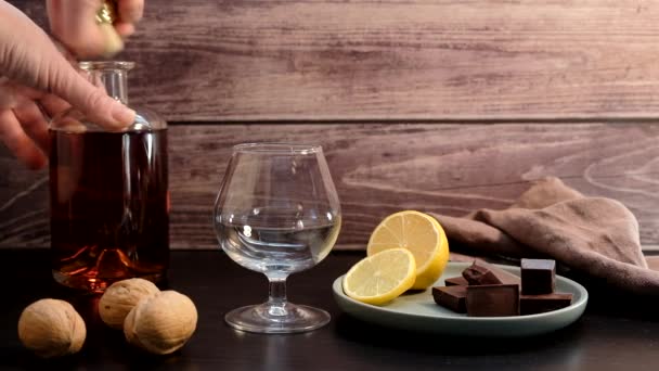 The hand pours cognac from the bottle into the wine glass. Next to it on a plate is a lemon and pieces of chocolate — Stock Video
