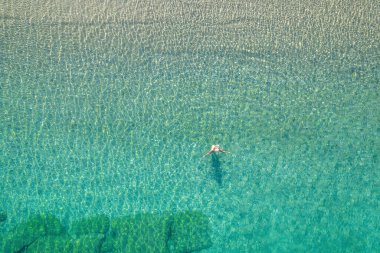Mayolu ve şapkalı genç bir kadın sahile yakın deniz dalgalarında yüzüyor. Yukarıdan bak. Üst, drone görünümü