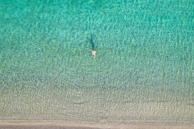 Mayolu ve şapkalı genç bir kadın sahile yakın deniz dalgalarında yüzüyor. Yukarıdan bak. Üst, drone görünümü