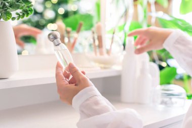 Makyaj aksesuarları ve masa aynasıyla eline parfüm süren bir kadın. Noel, ampuller, okaliptüs, köknar, bir vazoda çam dalları.