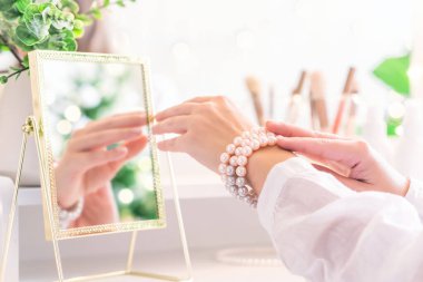 Kadın takı takıyor, eline inci bilezikler takıyor makyaj malzemeleriyle makyaj masasının yanında oturuyor. Kadın parti hazırlığı. Noel ağacının yansıması, masa aynasında ampuller..