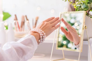 Kadın, makyaj malzemeleriyle birlikte sehpanın yanında duran inci bilezikleri veriyor. Noel ağacının yansıması, masa aynasında ampuller..