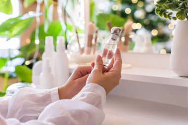 Makyaj aksesuarları ve masa aynasıyla eline parfüm süren bir kadın. Noel, ampuller, okaliptüs, köknar, bir vazoda çam dalları.