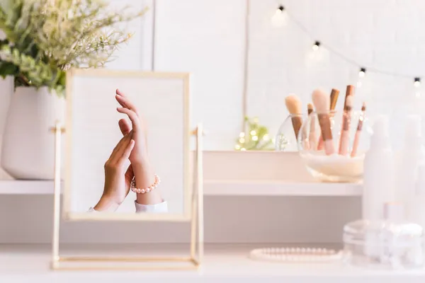 Reflection in table mirror of woman hand  in pearl bracelets on dressing table with make up accessories. Self-Care. White interior