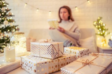 Noel ağacının yanındaki koltukta oturan ve el yapımı hediye kutularını masaya koyan kadın. Aile için hediyeler.